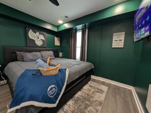 Quarto com cama, televisão fixada na parede, quadro com uma citação e janela com cortinas.