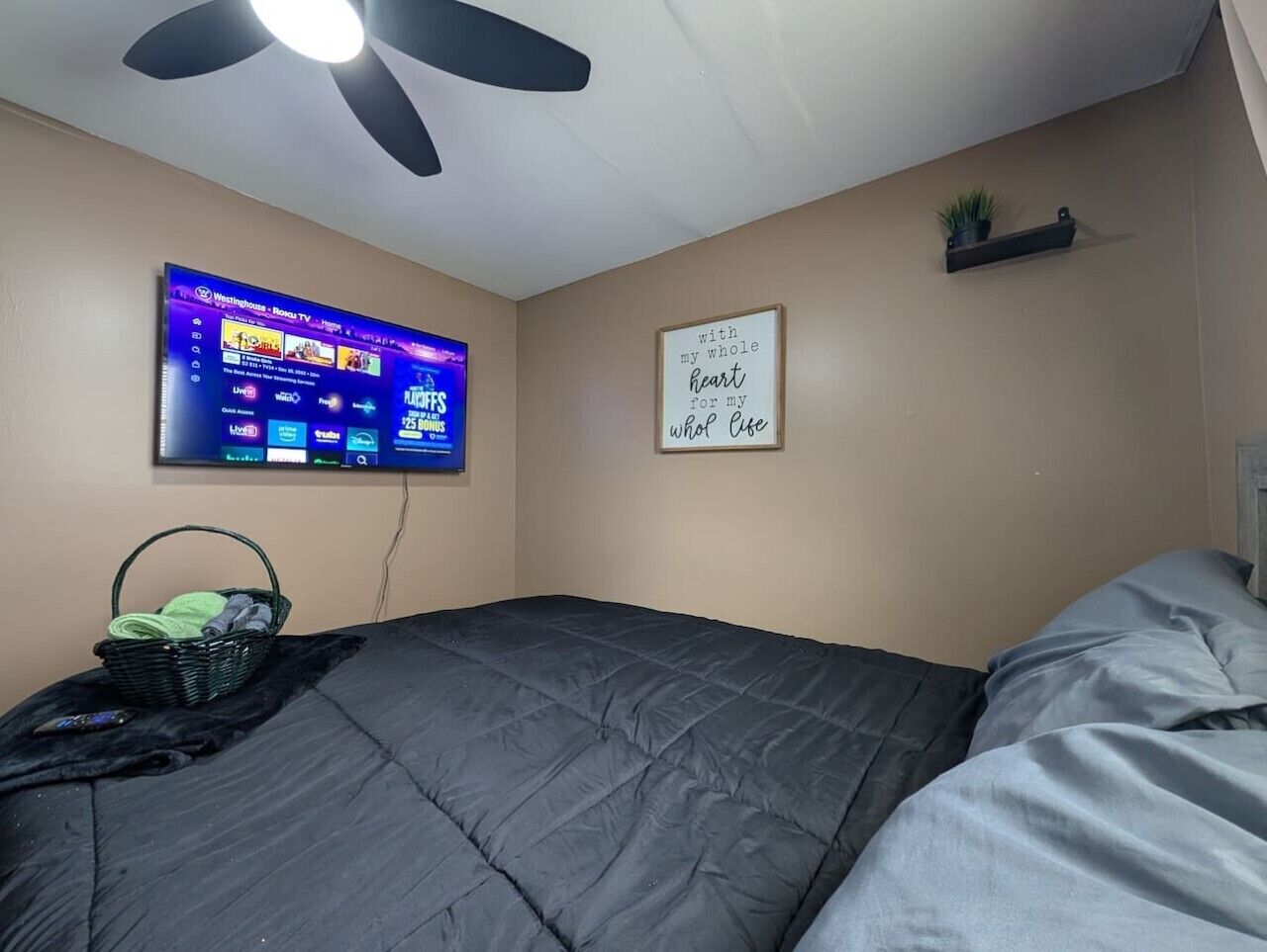 A bedroom with a bed, a ceiling fan, a mounted TV displaying a streaming service, a wall-mounted shelf with a plant, and a motivational quote on the wall.