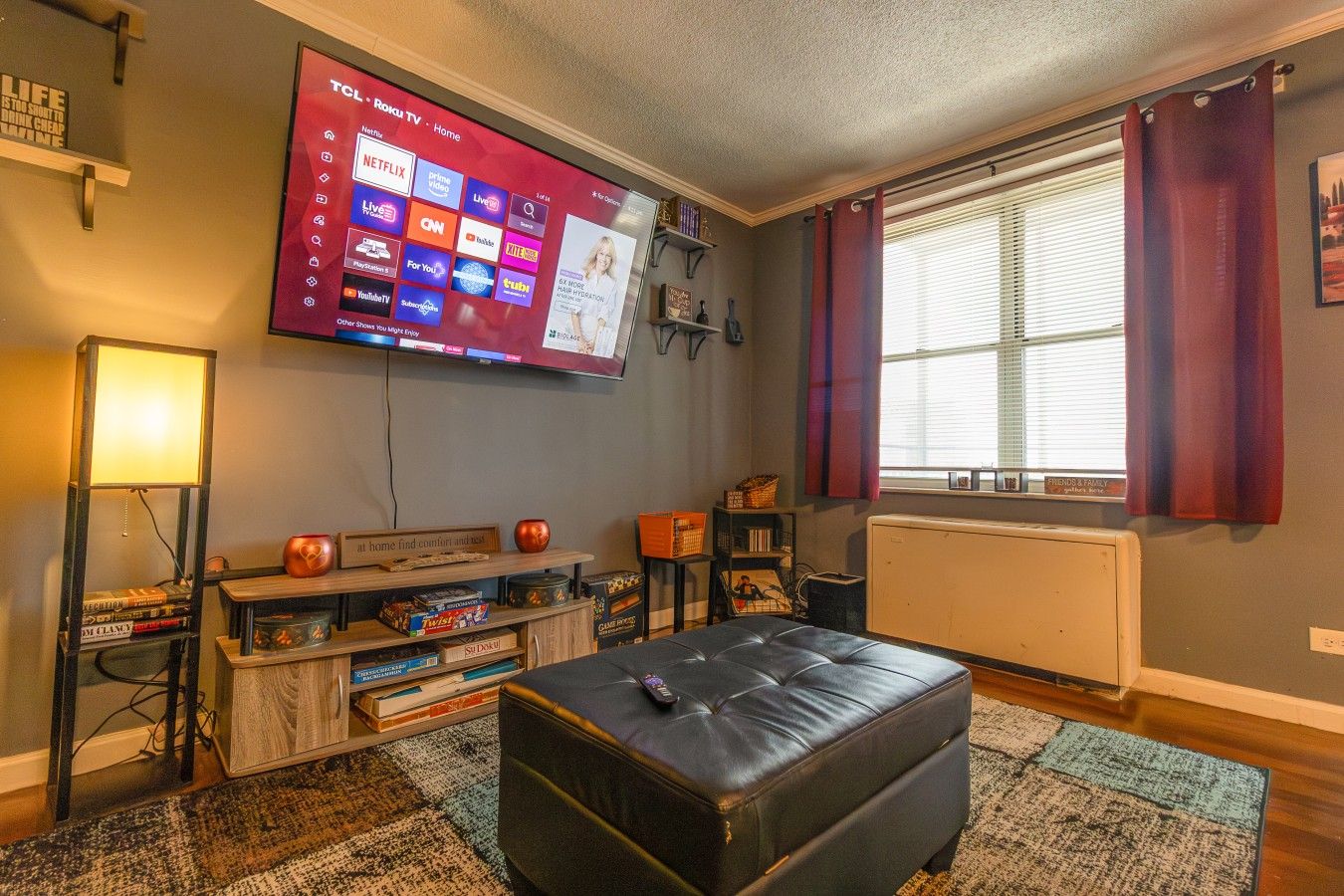 Un salon doté d’un téléviseur à écran plat fixé au mur, d’un pouf en cuir noir, d’une table basse en bois sur laquelle sont posés des jeux de société, et d’une fenêtre avec des rideaux.