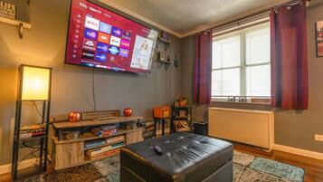 A living room with a flat-screen TV mounted on the wall, a black leather ottoman, a wooden coffee table with board games, and a window with curtains.