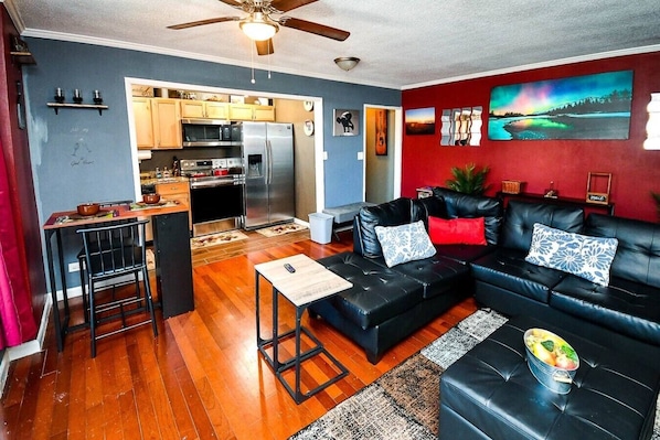 A living room with a black leather sofa, a dining area with a wooden table, and a kitchen with stainless steel appliances.