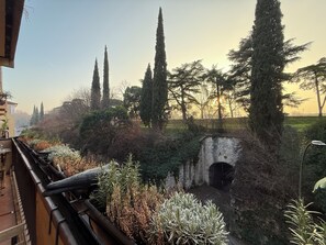 Een balkon met uitzicht op een stenen muur, hoge bomen en een weg in de verte.