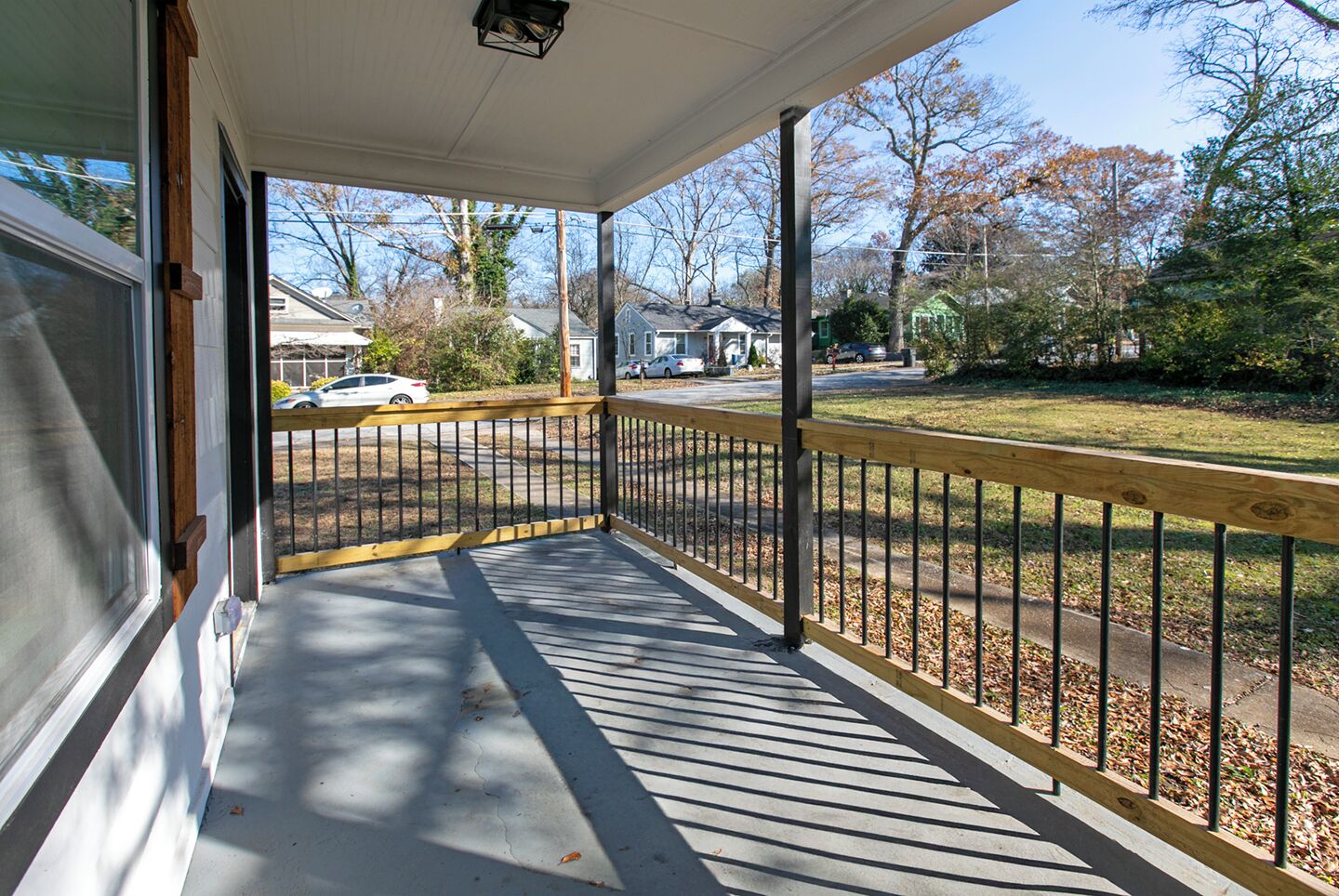 A covered porch with a wooden railing, a concrete floor, and a view of a residential street with houses and parked cars.
