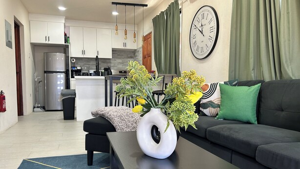 Una sala de estar moderna con un sofá, una mesa de centro con un florero, un reloj y una cocina al fondo.