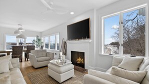 A modern living room with a fireplace, white sofas, a flat-screen TV, and large windows offering a view of the outdoors. - Family Waterfront Vacation Estate in New London, CT. (New London)