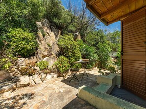 A stone patio with a table and chairs, surrounded by lush greenery and a rocky backdrop. - Casetta Nel Blu by Interhome (Villasimius)
