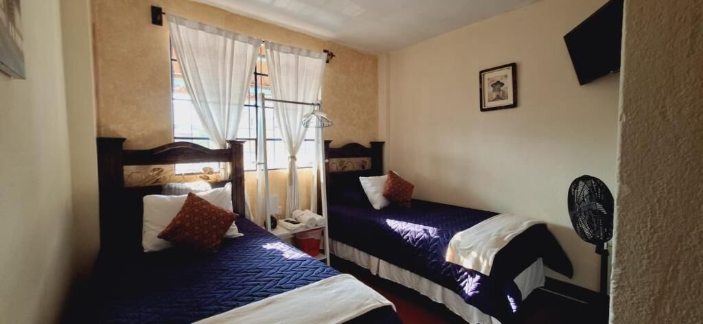 A room with two beds, a window with curtains, a bedside table, and a wall-mounted fan.