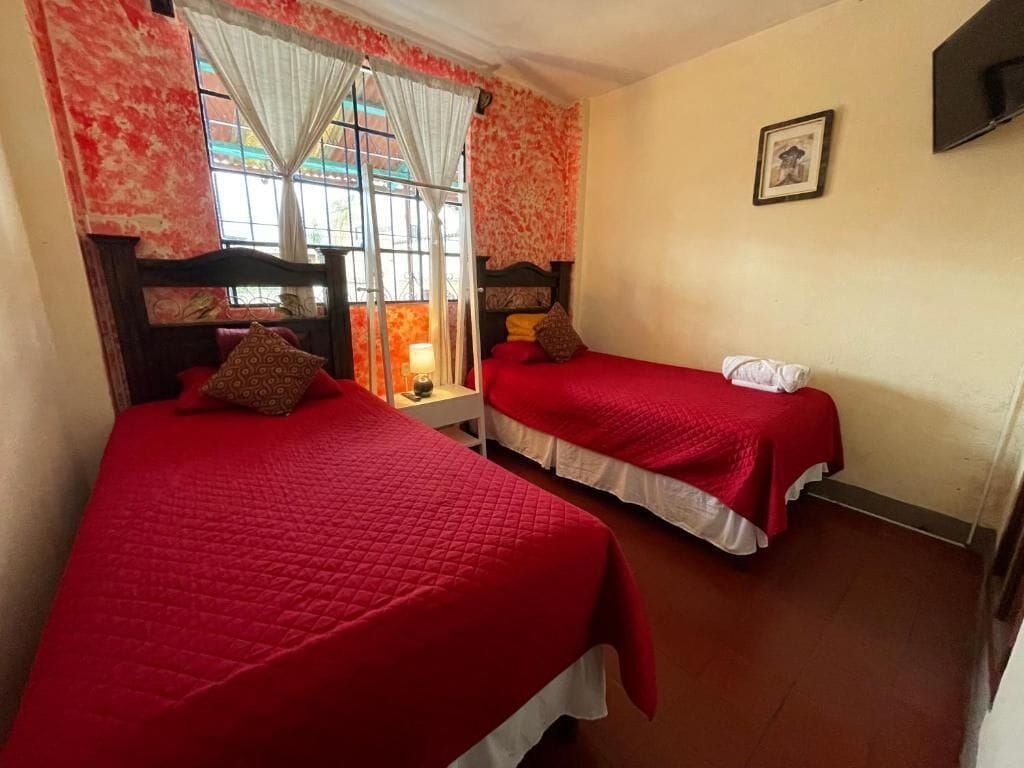 Habitació amb dos llits, cadascun amb una colcha vermella i llençols blancs. Hi ha una finestra amb cortines, una tauleta de nit amb una làmpada i un quadre al mur.