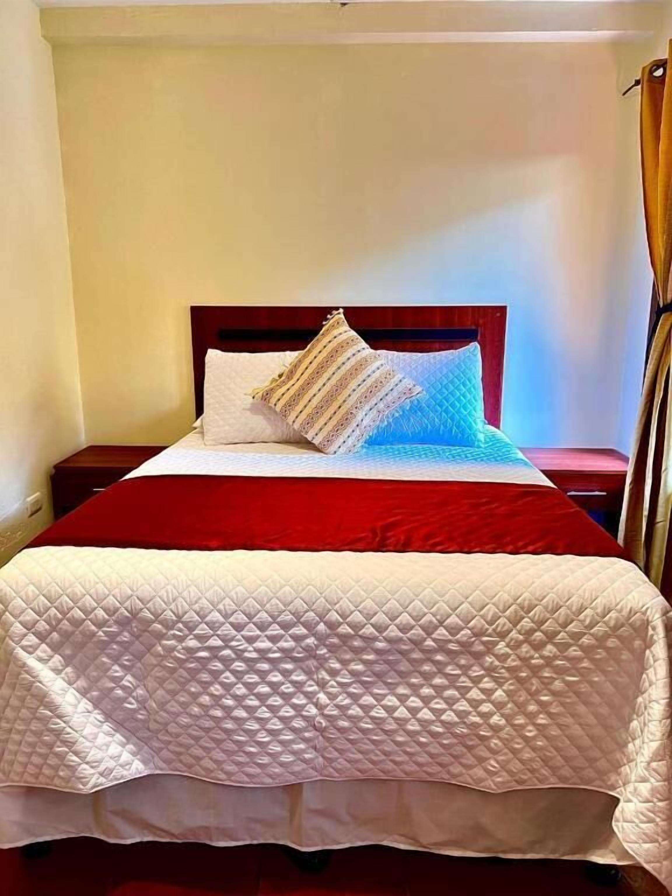 Una cama bien tendida con colcha roja y blanca, dos almohadas y cabecera de madera.