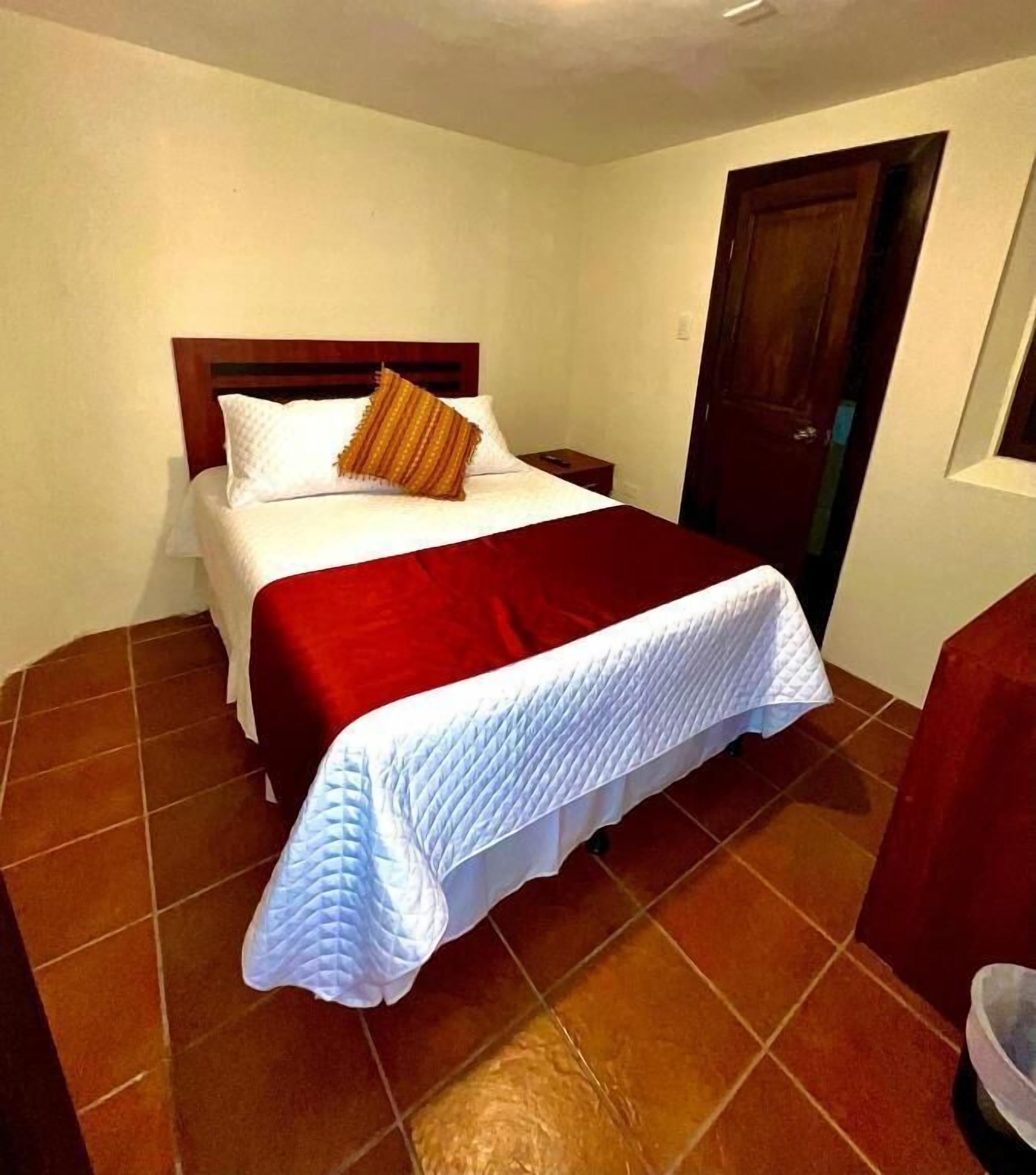 A hotel room with a bed, a wooden headboard, a red blanket, and a white quilted bedspread.