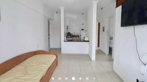 Ein Schlafzimmer mit Bett, einer kleinen Küchenzeile und einem an der Wand befestigten Fernseher.