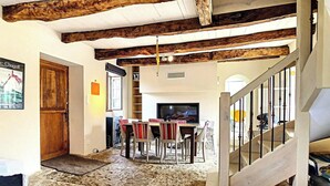 Un interior rústico con vigas de madera a la vista, una zona de comedor con sillas y una mesa, una escalera y un televisor.