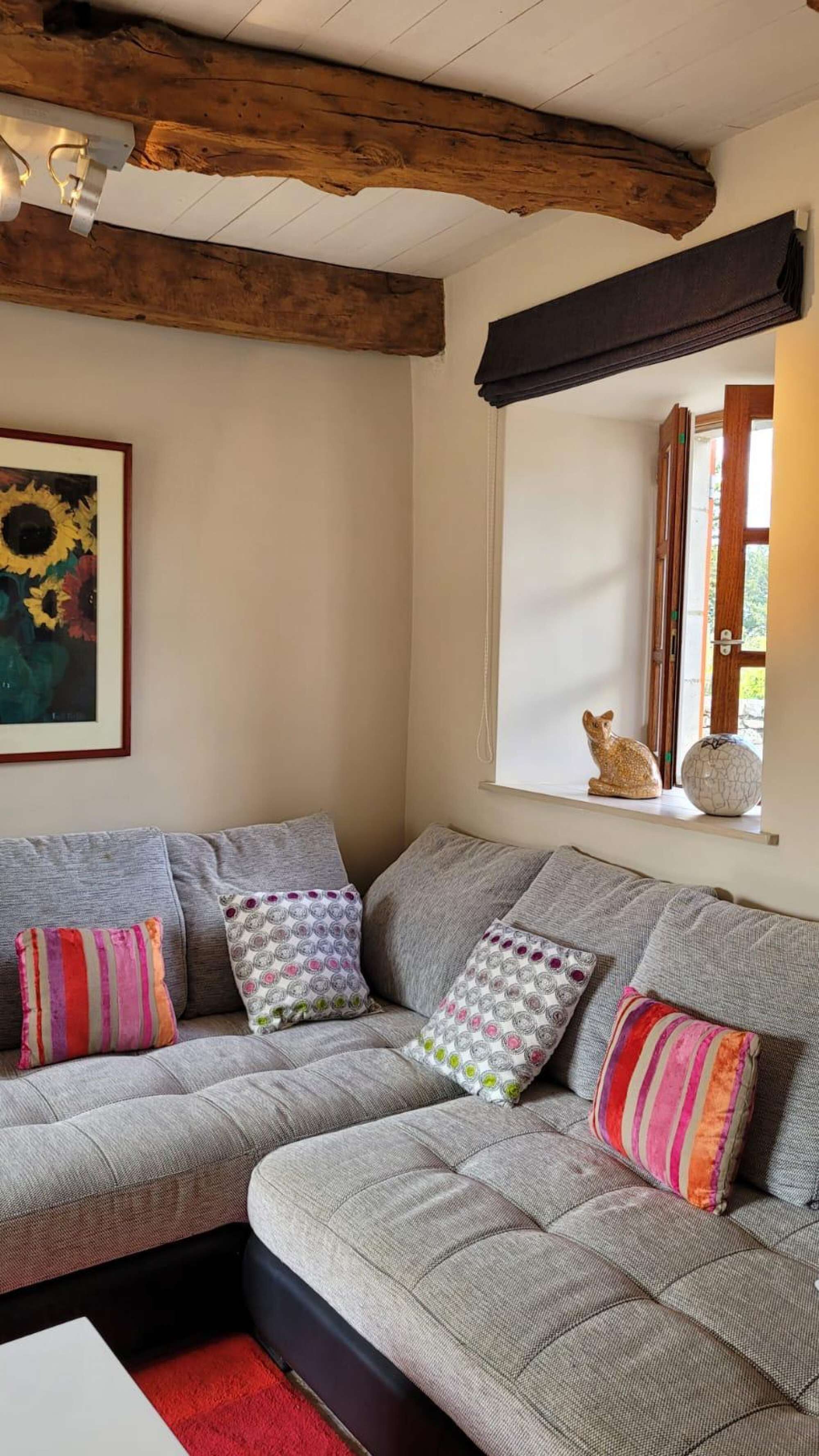 A cozy living room with a grey sectional sofa, colorful cushions, a framed sunflower painting, a cat figurine, and a window with a view.