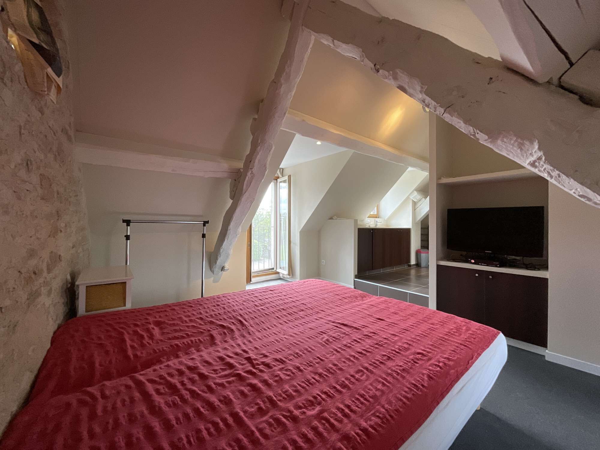 A bedroom with a red bedspread, a ceiling with exposed beams, a kitchenette, and a TV.
