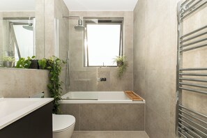 A modern bathroom with a bathtub, toilet, and shower area. There are plants on the windowsill and a towel rack. - Beautiful 1BR, Huge Terrace 17 mins central London - Pass the Keys (Surbiton)