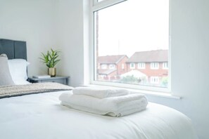 Un lit bien fait, avec des serviettes blanches pliées, une plante sur la table de chevet et une fenêtre donnant sur des maisons.