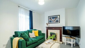 A living room with a green sofa, a flat-screen TV, a fireplace, and a painting on the wall. - Modern 3 Bedroom House | WA10 | Central St. Helens (St Helens)