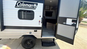 A Coleman RV with a sliding door open, revealing a compact interior with a bed, a small kitchenette, and a bathroom.