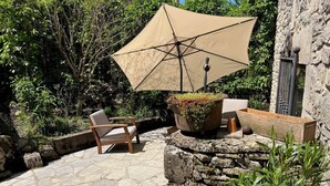 A patio with stone flooring, a wooden chair, a table, a beige umbrella, and a planter with green plants. - Lou Castel - Medieval castle (Le Clapier)