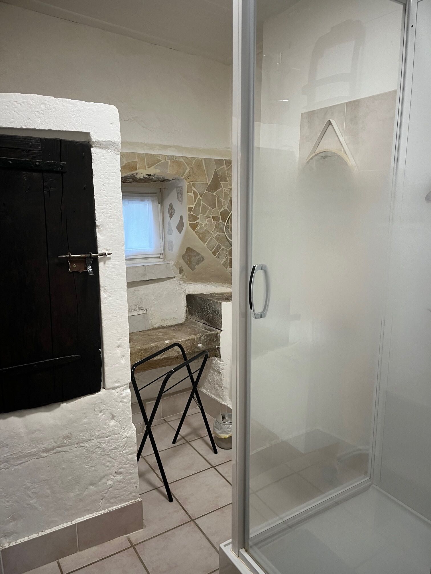 A bathroom with a glass shower door, a black door, a stone wall, and a small window.