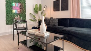 Un salon moderne avec une table basse en verre, un canapé foncé et un mur vert orné d'un néon.