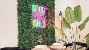 Un espace repas agrémenté d’un néon affichant le message « SEULEMENT les bonnes ondes », une table ronde en bois avec des assiettes et des verres, des chaises noires et une grande plante verte.