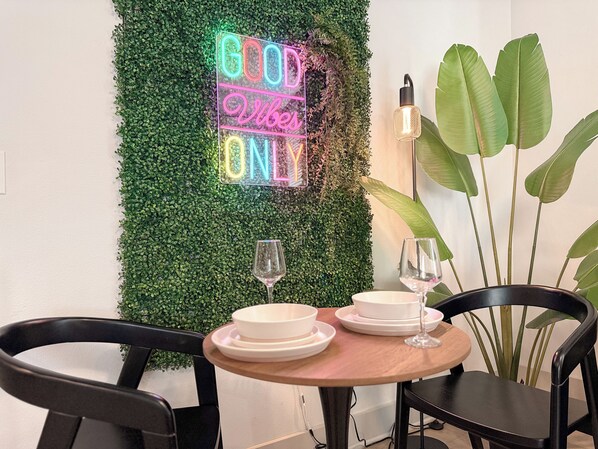 A dining area with a neon sign reading "GOOD Vibes Only", a round wooden table with plates and glasses, black chairs, and a large green plant.