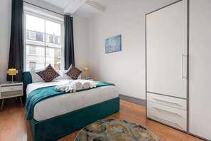A hotel room with a bed, a nightstand, a lamp, a wardrobe, and a view of the outside. - Stylish 2 BR Flat near Brick Lane, London Sleeps 4 (Greater London)