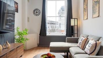 A modern living room with a sofa, a coffee table with fruit, a plant, and a wall clock.