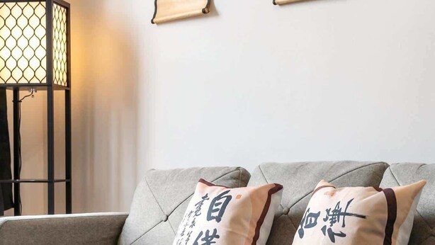 Een woonkamer met een grijze bank, twee kussens met kalligrafie, een ronde houten salontafel met fruit en een vloerlamp.