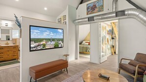 Een moderne woonkamer met een grote flatscreen-televisie, een houten bankje, een ronde salontafel en een stadsgezicht door het raam.