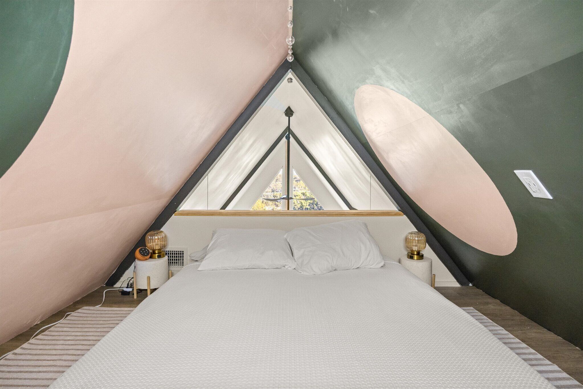 Een kamer met een driehoekige vorm, een bed, twee nachtkastjes met lampen, en een raam aan de bovenkant.