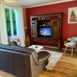 A living room with a brown leather sofa, a wooden entertainment unit with a TV, a colorful chair, and a window with curtains. - House with garden, close to shops, 4 km from the sea. Pets welcome. (Trégunc)
