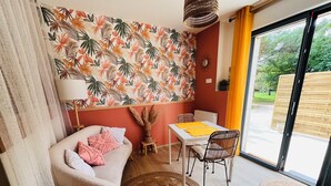 Un salon comprenant un canapé, une table à manger avec des chaises, un mur orné d’un motif, une fenêtre donnant sur un paysage verdoyant et un tabouret en osier.