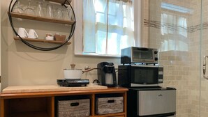 A compact kitchen with a microwave, coffee maker, and a pot on a portable stove. There are wooden drawers and a shelf with glassware.