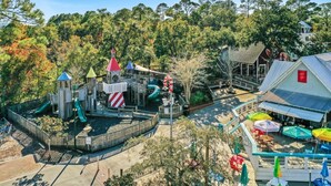 Ein Spielplatz mit Rutschen und Klettergerüsten, umgeben von Bäumen und einem Gebäude mit rotem Dach.