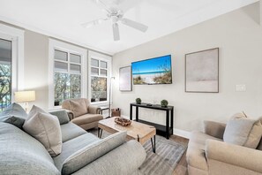 A living room with a beige sofa, a coffee table, and a ceiling fan. There are two windows with white frames and a view of trees outside. - Large private balcony! Can be a 2/2 or add Flip Flip for a 3/3 (Miramar Beach)