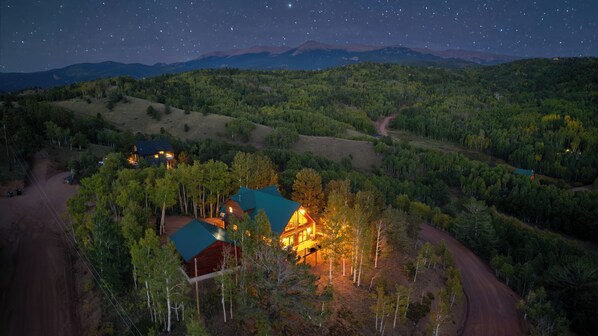 A remote cabin with a large wooden structure and a smaller attached building, surrounded by dense forest and a dirt road.