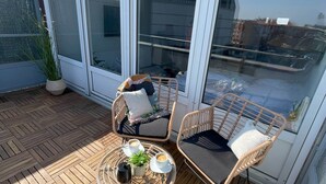 A rooftop terrace with a wooden floor, two wicker chairs, a small round table with a plant and a cup, and a large sliding glass door. - W&D Living-Tête de Fleur-Balcony above Schloßarkaden (Brunswick)