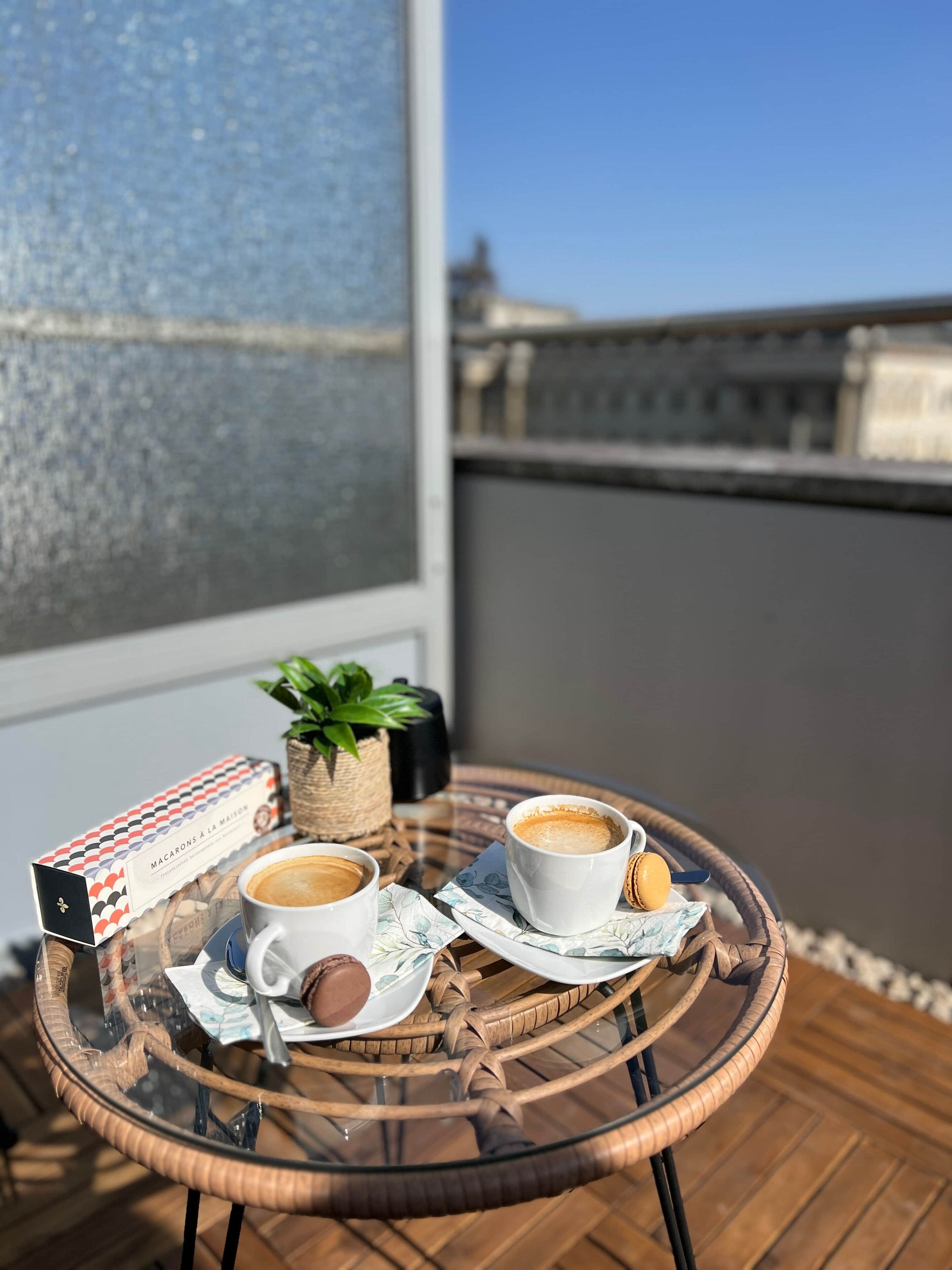 A glass table with two cups of coffee, a box of macarons, a small plant, and a wicker basket on a balcony.