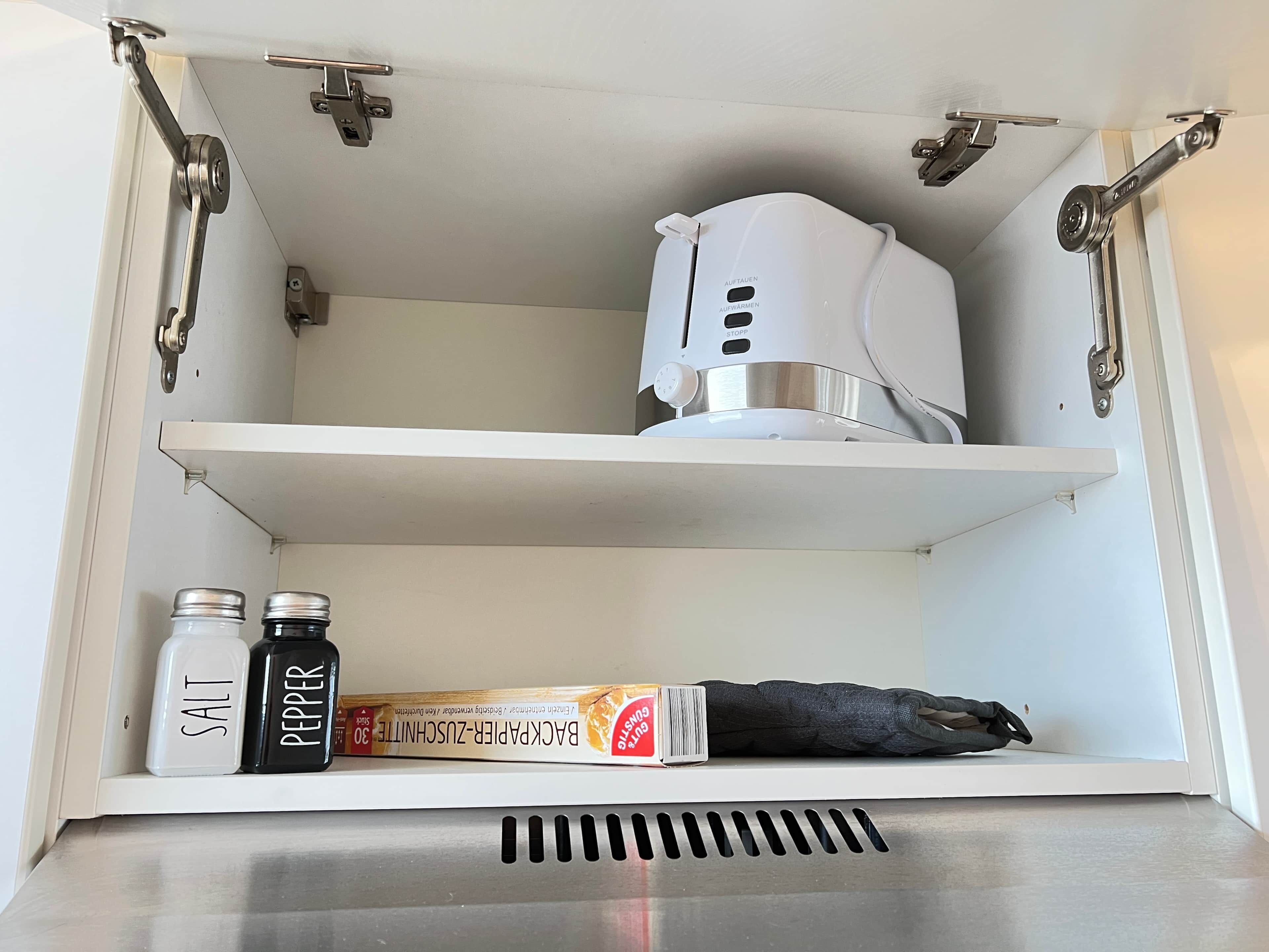 A kitchen cabinet with a white appliance on the top shelf, salt and pepper shakers on the bottom shelf, and a cookbook.