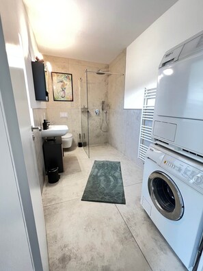 Casa de banho compacta com chuveiro, sanita, lavatório e máquina de lavar roupa frontal.