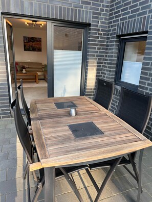 Mesa de pátio com cadeiras, cafeteira e porta de vidro deslizante que dá para uma área de estar interna com sofá e um quadro.