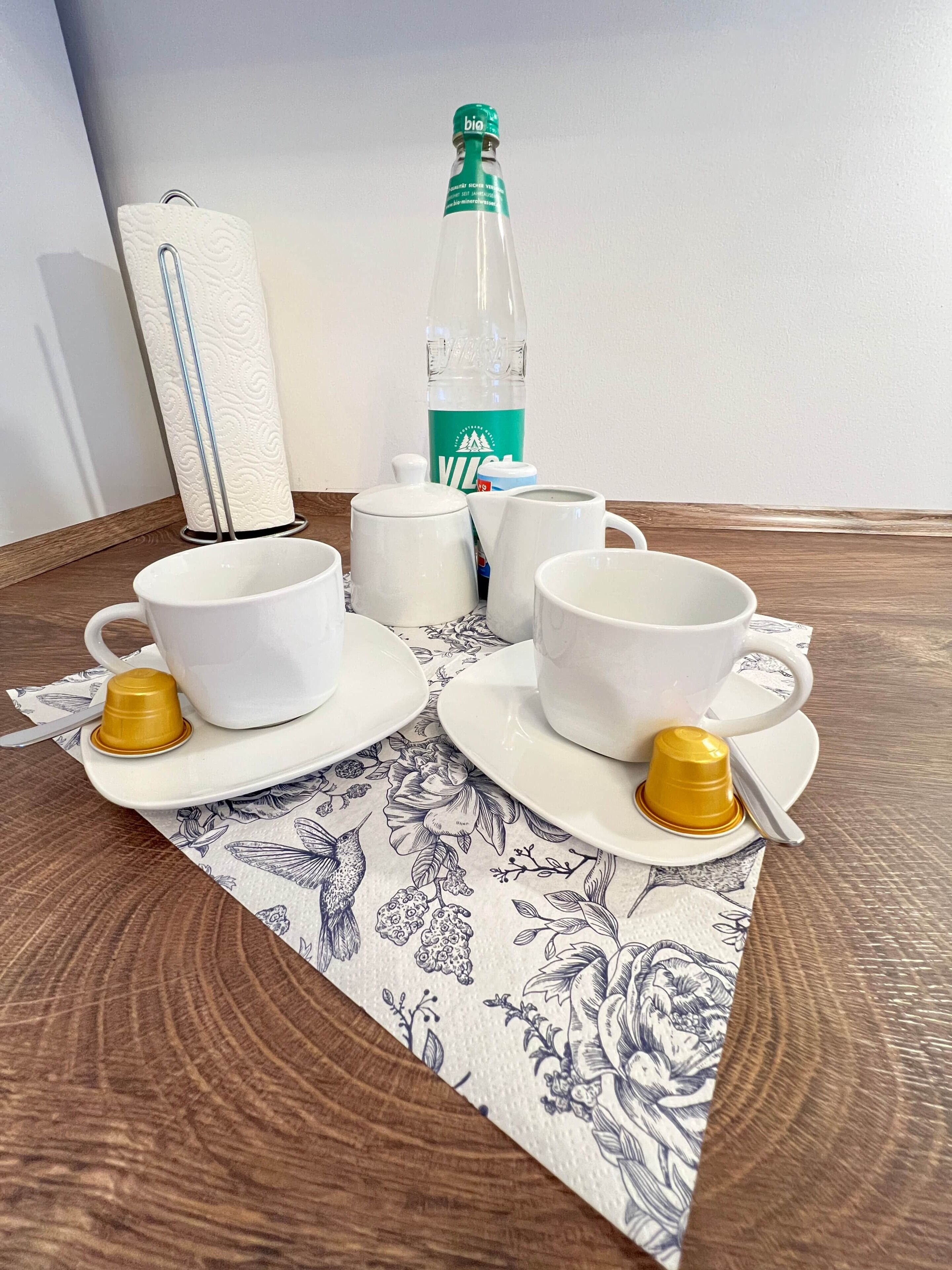 Ein Tisch, gedeckt mit weißen Tassen und Untertassen, einer Flasche und einer Serviettenrolle.
