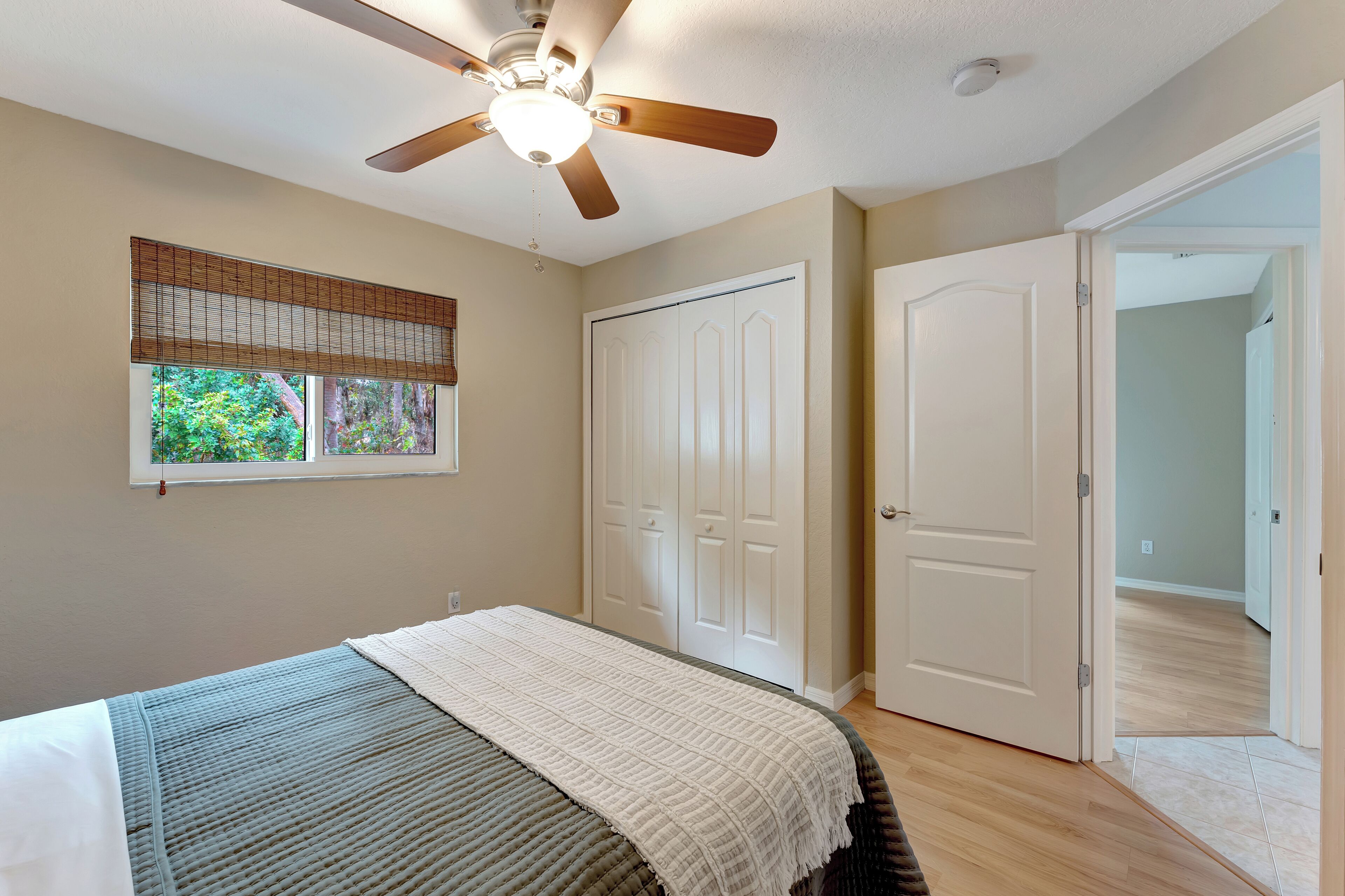 A bedroom with a bed, a ceiling fan, a window with blinds, and a door leading to another room.