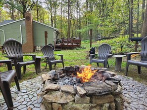 A stone fire pit surrounded by Adirondack chairs, a wooden deck, and a cabin in a wooded area. - Fern House: Your Quiet Escape in the Woods (Honey Brook)