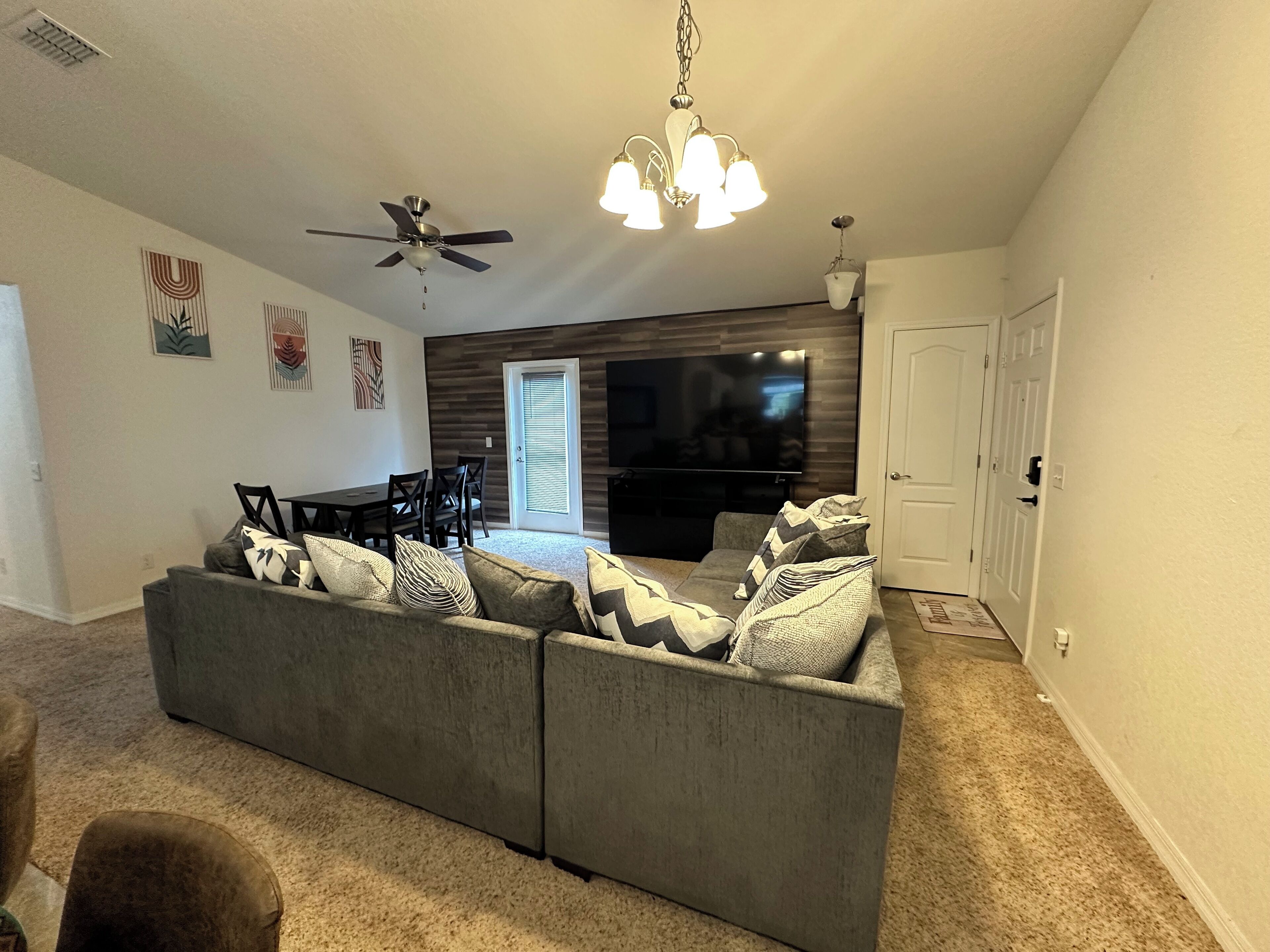 A living room with a gray sectional sofa, a ceiling fan, a chandelier, and a dining area with a table and chairs.