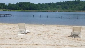 Una playa con dos sillas de playa vacías orientadas a un lago tranquilo, rodeada por una cerca de malla y un muelle al fondo.