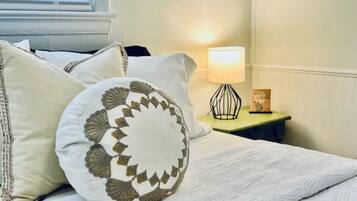 Una cama bien hecha con ropa blanca y almohadones decorativos, una lámpara de noche y una mesita con una vela y un libro.