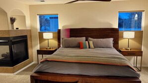 Une chambre à coucher avec un lit en bois, deux tables de chevet avec des lampes, une cheminée et une vue sur les arbres à travers les fenêtres.
